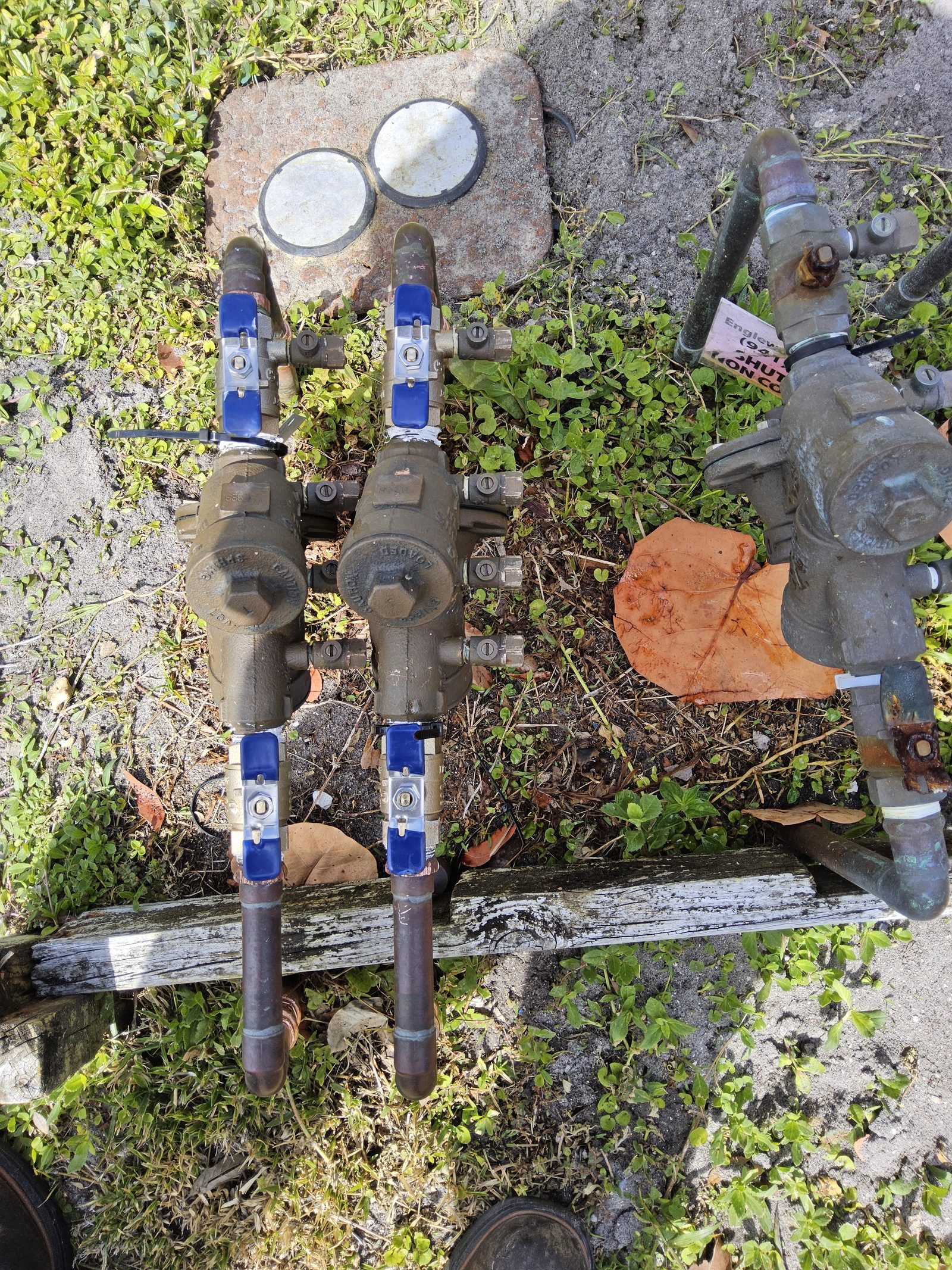 Overhead view of a residential property with a corroded older backflow assembly in the background and a newer dual-check Febco assembly with blue ball-valve handles in the foreground after replacement.
