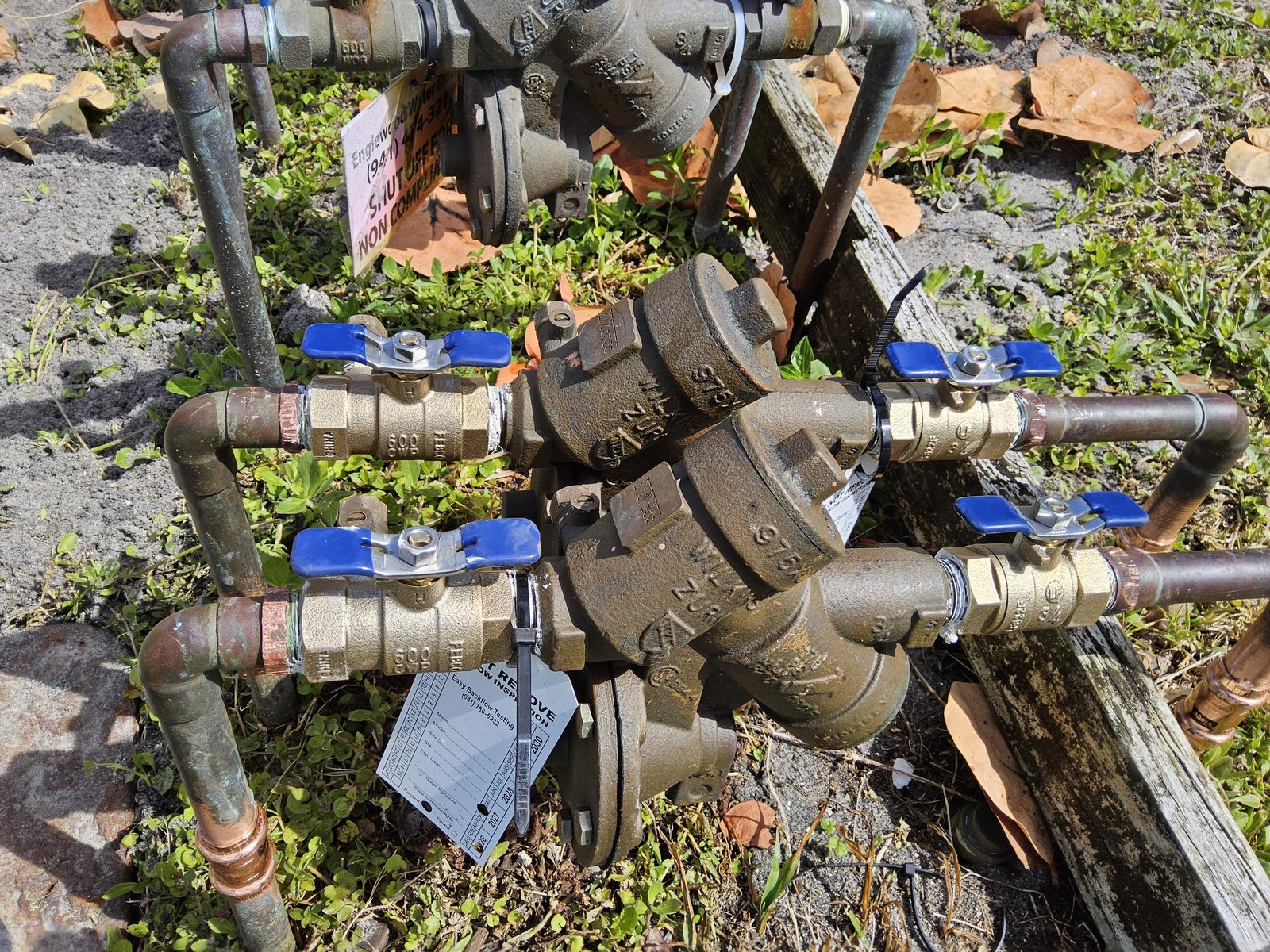 Dual Wilkins Zurn 975XL2 RPZ backflow preventer with a fresh passing inspection tag, restored to compliance after an Englewood Water District shut-off notice for non-compliance visible in the background.